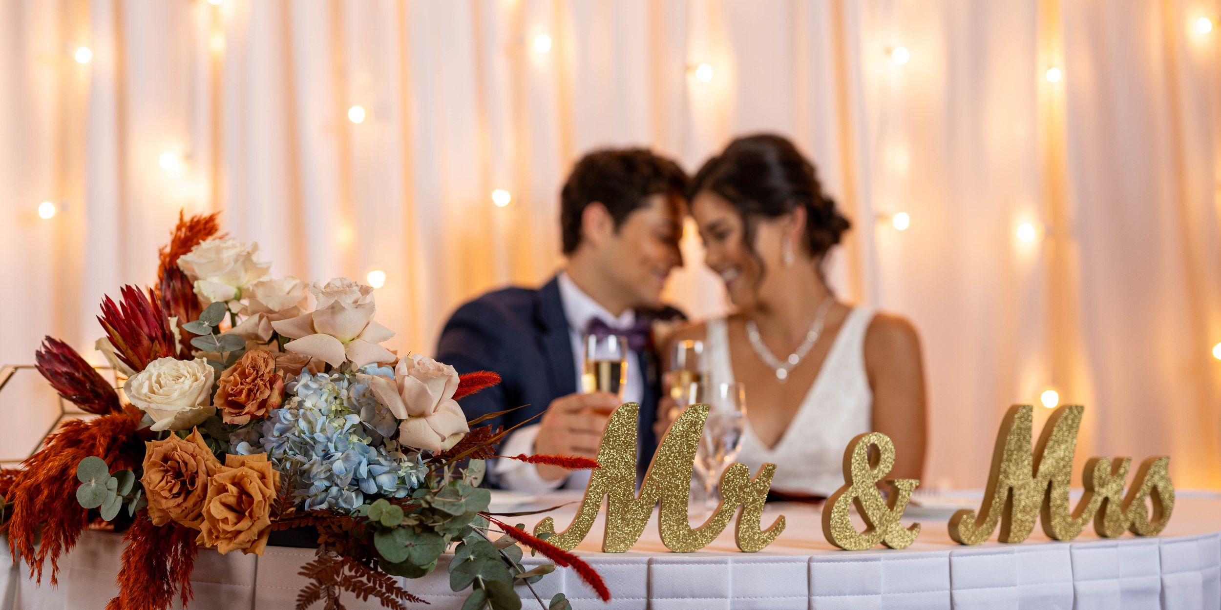 A Bride And Groom Sitting At A Table With Flowers