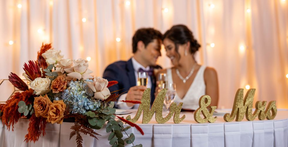 A Bride And Groom Sitting At A Table With Flowers